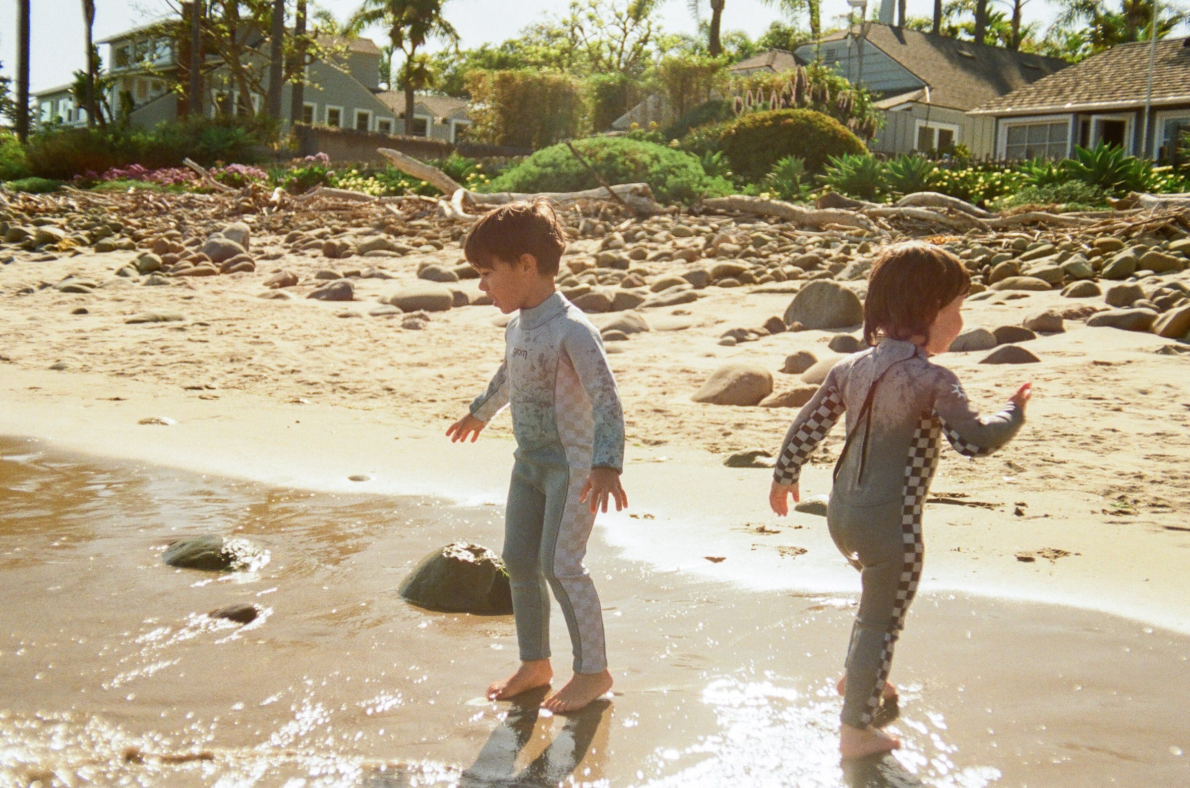 Two toddlers in Le Grom checkerboard wetsuits walking a sunlit California cobble beach