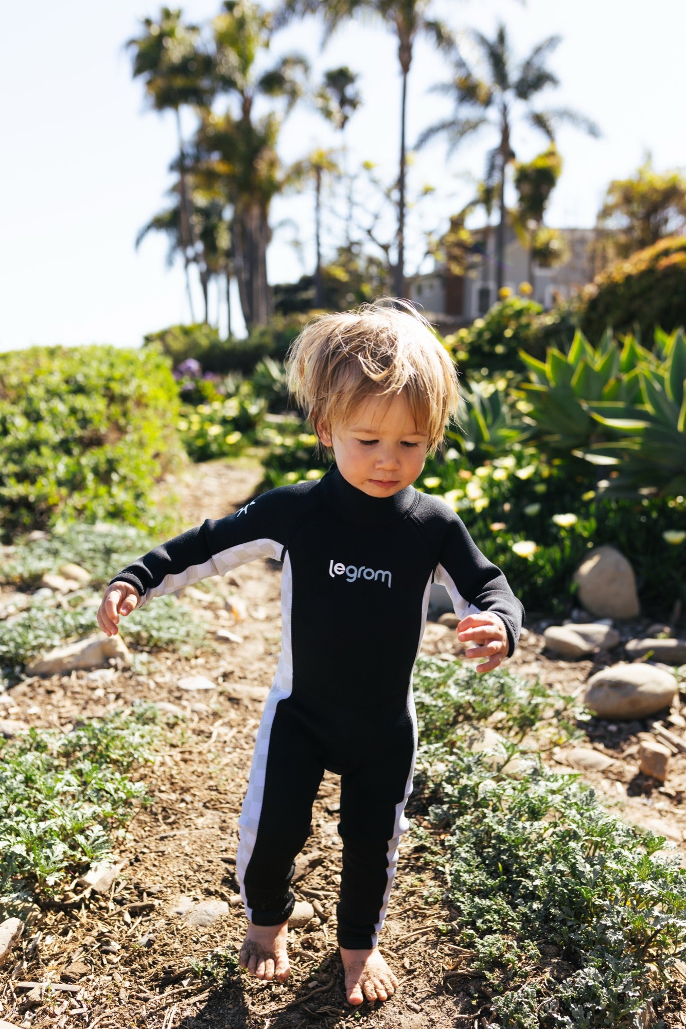 A toddler in a Le Grom wetsuit walking a sunlit garden path, palms overhead